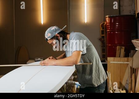 Side view of male shaper using measure tool while working with surfboard in workshop Stock Photo