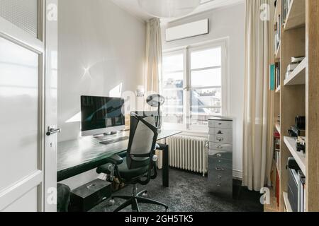 Comfortable armchair placed near table with computer monitor in modern workplace with shelves in contemporary apartment Stock Photo