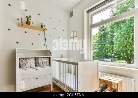 White crib placed between cabinet and bookshelf near window in sunlit nursery at home Stock Photo