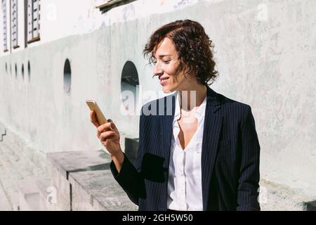 Positive adult female executive worker wearing classy outfit standing browsing on cellphone in sunny day Stock Photo