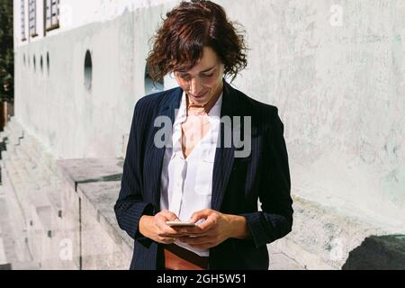 Positive adult female executive worker wearing classy outfit standing browsing on cellphone in sunny day Stock Photo