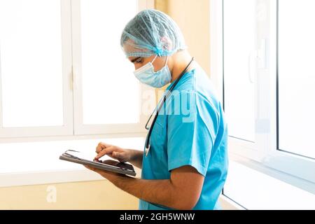 Male medic in uniform and with stethoscope standing in hospital and surfing Internet on tablet Stock Photo