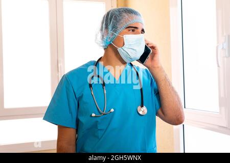 Male medic in uniform and with stethoscope standing in hospital and talking on smartphone Stock Photo