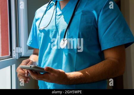 Unrecognizable crop doctor in uniform using mobile phone while standing in corridor near window in hospital Stock Photo