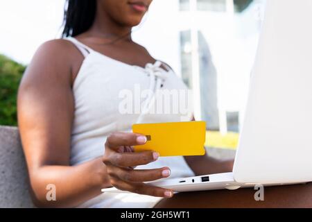 Low angle of anonymous African American female making payment with plastic card while using laptop during online shopping in Barcelona street Stock Photo