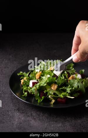 Crop anonymous cook with kitchen tongs serving appetizing beetroot salad with Tofu cheese and lettuce leaves on plate on dark table in studio Stock Photo