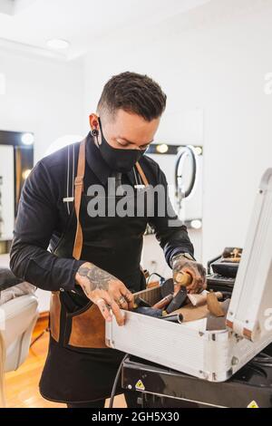 Tattooed man in mask picking various cosmetic supplies from suitcase while working in beauty salon during pandemic Stock Photo
