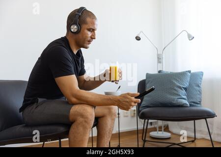 Side view from above of male in headphones watching video on tablet while drinking orange juice and sitting on chair in morning at home Stock Photo