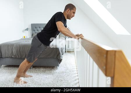 Side view of male doing push ups near wooden railing while exercising in morning in bedroom Stock Photo