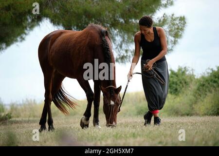 Full body of ethnic female stroking obedient brown stallion with bridle while walking together on green meadow with trees in summer countryside Stock Photo