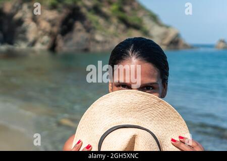 Ethnic female tourist hiding face behind straw hat while looking at camera against ocean and mountain in sunlight Stock Photo