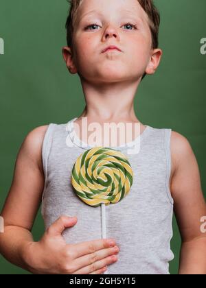 Kid with sweet swirl lollipop towards camera on green background in studio Stock Photo