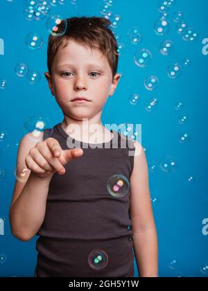 Astonished preteen boy showing touching gesture with index finger in air in studio with flying soap bubbles on blue background Stock Photo