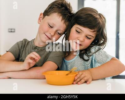 Adorable twin sister and brother sitting at table with bowl of food during lunch at home Stock Photo