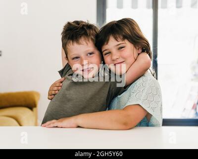 Cute content twins embracing tenderly and sitting together in living room at home looking at camera Stock Photo