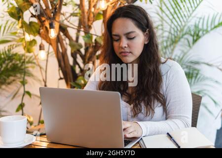 Focused young Latin American female student reading information on laptop screen while preparing for university exam in cozy cafe Stock Photo