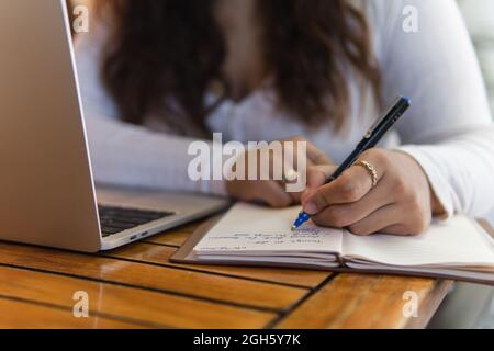 Cropped unrecognizable female student taking notes in planner while working on laptop preparing for university exam in cozy cafe Stock Photo