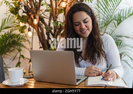 Focused young Latin American female student taking notes in planner while working on laptop preparing for university exam in cozy cafe Stock Photo