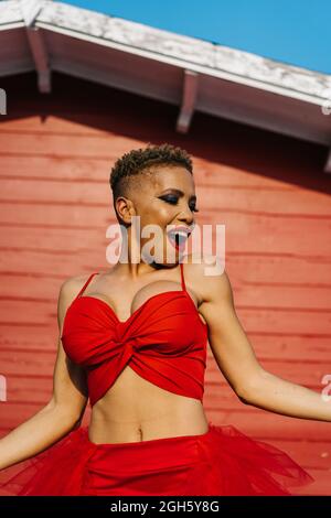 Trendy African American female with red lips and short hair dancing while shouting against house wall Stock Photo