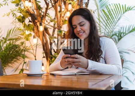 Young ethnic female messaging on mobile phone with notepad while sitting at table with cup of coffee and notebook in cafeteria Stock Photo