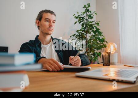 College student preparing for pass exam at home. Young man making notes while online learning Stock Photo