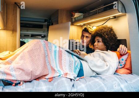 Happy romantic multiracial couple cuddling and enjoying time together while lying on bed and watching movie on laptop during travel in camper van Stock Photo
