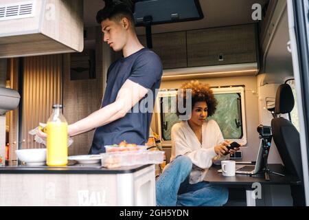 Young guy preparing healthy breakfast while African American girlfriend using gadgets during summer journey together in camper van Stock Photo