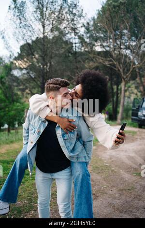 Young man giving cheerful ethnic girlfriend piggyback ride against camper and Thai Ridgeback on pathway Stock Photo