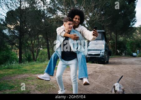 Smiling black Woman riding piggyback on boyfriend while taking self portrait on cellphone against dog and caravan in camp Stock Photo
