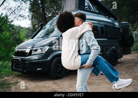 Cheerful young African American Woman laughing happily and embracing boyfriend while having fun together near camper van parked in green forest during Stock Photo