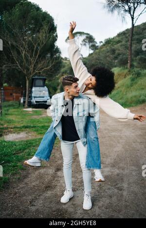 Young man giving cheerful ethnic girlfriend piggyback ride while looking away against camper and Thai Ridgeback on pathway Stock Photo