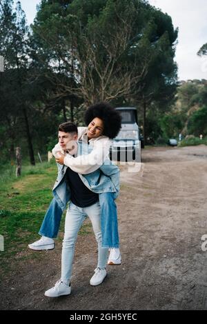 Young man giving cheerful ethnic girlfriend piggyback ride while looking away against camper and Thai Ridgeback on pathway Stock Photo