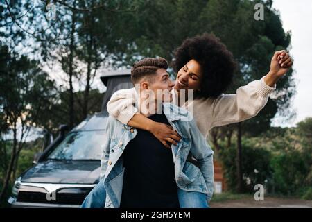 Young man giving cheerful ethnic girlfriend piggyback ride against camper and Thai Ridgeback on pathway Stock Photo