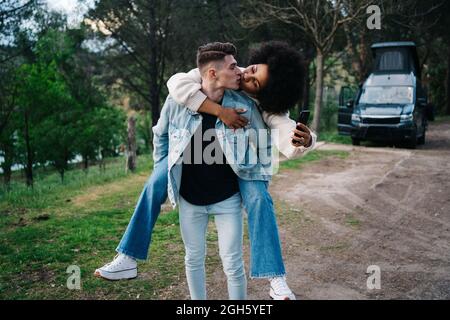 Young man giving cheerful ethnic girlfriend piggyback ride against camper and Thai Ridgeback on pathway Stock Photo