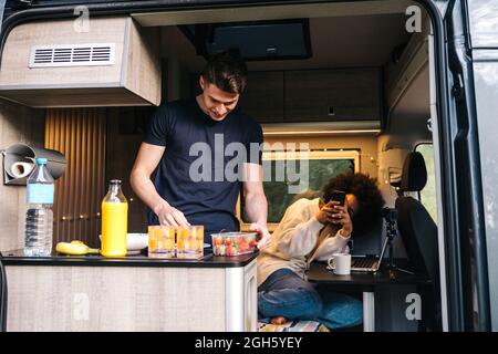 Young African American Woman with smartphone taking photo of boyfriend preparing healthy meal inside camper van during summer holidays together Stock Photo