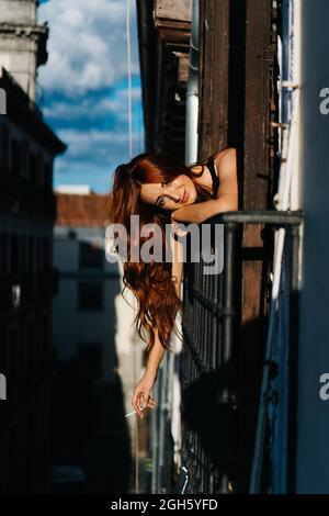 Delighted redhead female with cigarette sticking out of balcony and leaning on metal railing at sunset and looking at camera Stock Photo