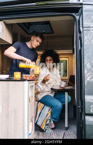 Young man serving natural juice to cheerful black girlfriend while enjoying breakfast together inside camper trailer during summer holidays Stock Photo