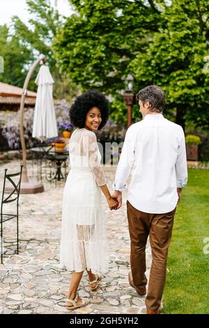 Back view of man holding hand of loving Woman looking over shoulder at camera during stroll in summer park Stock Photo