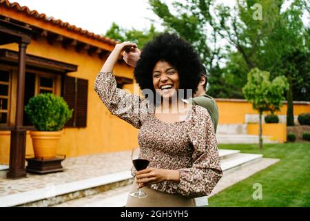 Content black Woman with glass of red wine holding hand of man and dancing in yard near cottage while having fun Stock Photo