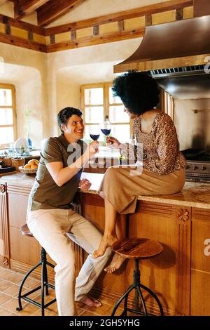 Delighted black Woman sitting on counter and man sitting on stool in kitchen clinking glasses with alcohol drink while celebrating event at home and l Stock Photo