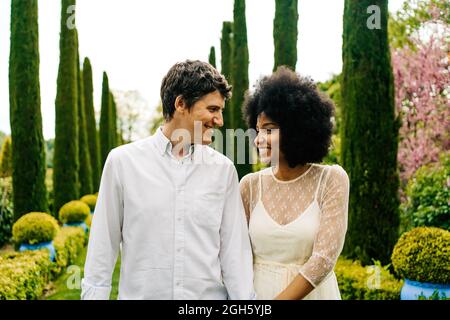 Cheerful multiracial couple holding hands and walking in garden while looking at each other Stock Photo