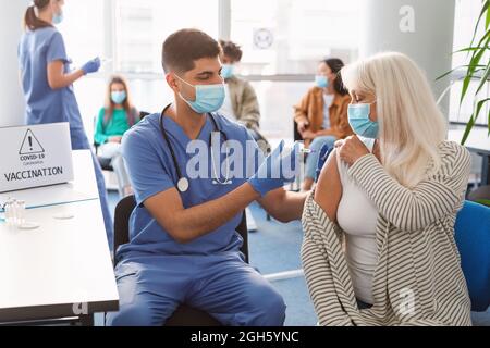 Mature Lady Receiving Coronavirus Vaccine Injection Sitting With Doctor Indoor Stock Photo