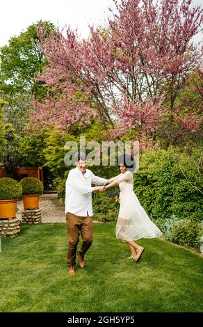 Optimistic multiracial couple holding hands and dancing on lawn while having fun in summer garden Stock Photo