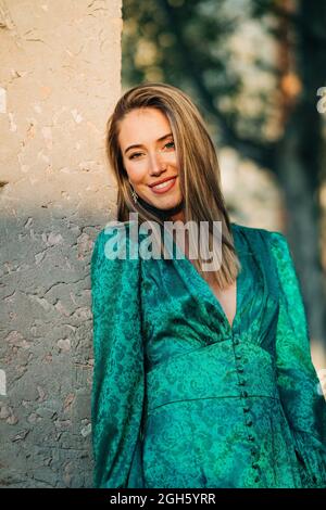 Elegant lady in hat and leather jacket shooting on camera and standing on footpath between murk alley of high walls and woods Stock Photo