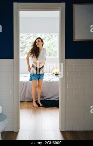 Charming slim female putting on denim shorts while standing in bedroom at home Stock Photo