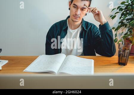Pensive student making notes from laptop. Young man studying at home and prepare for pass exam Stock Photo