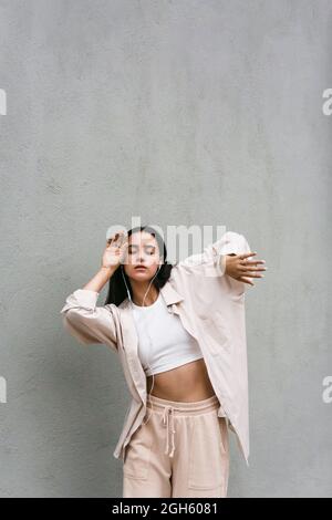 Carefree female dancer listening to music in earphones and dancing with closed eyes while enjoying songs on background of gray wall in city Stock Photo