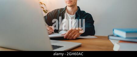 College student preparing for pass exam at home. Young man making notes while online learning. Wide image Stock Photo