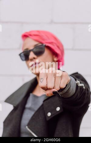 Young trendy female in stylish outfit and sunglasses showing fist with small  tattoo on finger while standing near white wall on street Stock Photo -  Alamy