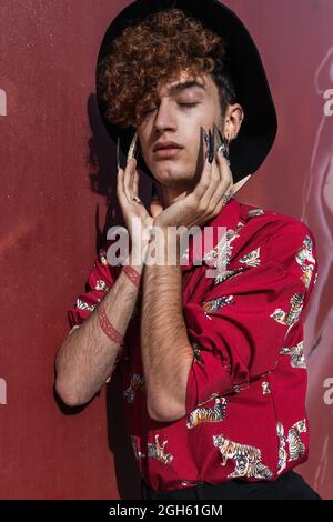 Dreamy young feminine gay with closed eyes and tattoo in stylish apparel touching face in sunlight Stock Photo